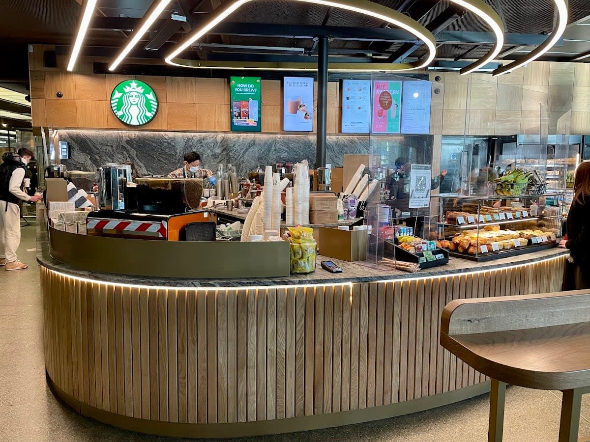 Early morning commuters blur past a Starbucks counter bathed in warm overhead light, their rolling luggage and glowing phone screens contrasting against the cool pre-dawn grey filtering through the station's glass canopy.